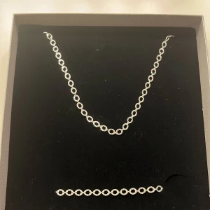 Silverfärgat halsband och armband set - Snyggt set med halsband och armband i silverfärg. Båda har en kedja med ovala, glittriga länkar som ger en stilren och modern look. Perfekt för dig som gillar minimalistiska smycken med det lilla extra. Dem är från guldfynd och är aldrig använda. Originalpris är 600