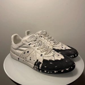 Maison Margiela svartvita gats - Maison Margiela sneakers i canvas med unik svart och vit färgskvätt-design. Klassisk låg modell med snörning, rund tå och platt sula. Märkets ikoniska nummeretikett på plösen. Skorna har en konstnärlig vibe och sticker ut med sitt handmålade utseende. Passar 45-46. Dustbags och kvitto finns, skriv gärna för frågor.