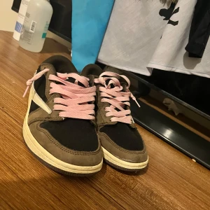 Nike Air Jordan 1 Low Travis Scott - Säljer ett par Nike Air Jordan 1 Low Travis Scott med bruna och svarta paneler i mocka och skinn, vit omvänd swoosh och rosa snören. Röd broderad logga på hälen. 