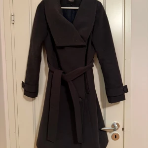 Grå kappa från Soaked in Luxury XS - Stilren mörkgrå kappa från Soaked in Luxury i storlek XS. Kappan har bred krage, knytskärp i midjan och dekorativa knappar framtill. Fickor i sidorna. Långa ärmar med snygga detaljer vid ärmslut. Perfekt för höst och vinter när du vill vara både varm och snygg. Väldigt fint skick.