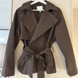 Brun trenchcoat från H&M med bälte - Snygg brun kort trenchcoat från H&M ❤️ Populär och helt slutsåld!! Använd en gång 