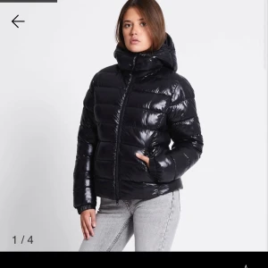 Svart glansig pufferjacka med huva - Säljer en svart glansig pufferjacka med huva och dragkedja. Jackan har en quiltad look med breda kanaler och är långärmad. Perfekt för kalla dagar när du vill hålla stilen och värmen. Snygg och enkel att matcha med jeans eller andra byxor.