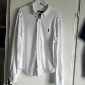 Vit långärmad skjorta från Polo Ralph Lauren - Klassisk vit skjorta från Polo Ralph Lauren med broderad marinblå logga på bröstet. Skjortan har lång ärm, knappar framtill och är gjord i mjuk bomull. Perfekt för en clean och stilren look. Har inte använts mycket. Ny pris 1095kr
