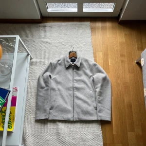 Ljusgrå ulljacka från H&M - Säljer en stilren ljusgrå jacka från H&M i ullblandning. Jackan har klassisk krage, dragkedja framtill och två snedställda fickor. Perfekt för dig som gillar enkel och clean stil. Passar till många olika outfits.