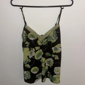 Blommig linnetopp med spetskant - Snygg linnetopp med grönt och vitt blommönster på svart botten. Toppen har tunna axelband och en v-ringad hals med grön spetskant som ger en extra detalj. Perfekt för dig som gillar en feminin och trendig stil.