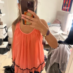 Persikofärgad blus  - Supersöt persikofärgad kortklänning med svarta vågiga kanter längst ner. Klänningen är ärmlös med rund halsringning och har en luftig, flowy passform som är perfekt för varma dagar.