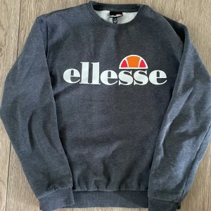 Mörkgrå sweatshirt från Ellesse - Mörkgrå sweatshirt från Ellesse med stort vitt logotryck och färgglad detalj på bröstet. Klassisk rund halsringning och ribbade muddar. Tillverkad i mjuk bomullsblandning för extra komfort. Perfekt för en avslappnad och sportig stil.