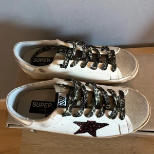 Golden Goose sneakers med glitterstjärna - Snygga Golden Goose sneakers i vitt skinn med silvriga detaljer och beige mocka framtill. Skorna har leopardmönstrade snören och en mörkröd glittrig stjärna på sidan. Perfekta för dig som vill sticka ut med coola detaljer och trendig design.