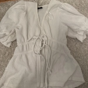 Vit topp från gina - Superfin vit topp med puffiga korta ärmar och knytband i midjan. Toppen  har en strukturerad, lite krispig bomullskänsla och v-ringad hals. Perfekt för varma dagar när du vill känna dig både söt och bekväm.