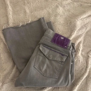 Grå bootcut jeans med lila detaljer - Snygga grå jeans från dvb med bootcut passform och råa kanter nertill. Byxorna har en unik lila patch och dragkedjedetalj på bakfickan. Perfekta för dig som vill sticka ut med coola detaljer och en klassisk siluett.