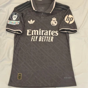 Real Madrid x Adidas grå matchtröja - Snygg grå Real Madrid fotbollströja från Adidas med broderade klubbmärken, HP-logga och Champions League 15-patch på ärmen. Tröjan har korta ärmar, krage med knappar och subtilt mönster i tyget. Denna tröjan är samma som spelarna använder(player version). Tröjan är från 2025 och är i extremt bra skick. Säljs billigt.