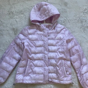 Ljuslila pufferjacka från Levi's L - Snygg ljuslila pufferjacka från Levi's med huva och dragkedja. Jackan har quiltad design, två fickor med dragkedja och justerbar nederkant. Perfekt för kalla dagar när du vill vara både varm och trendig.