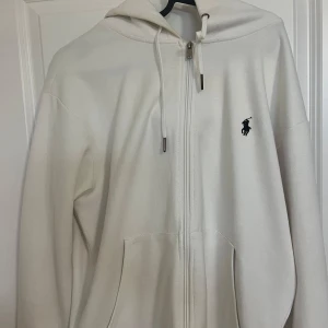 Vit hoodie från Polo Ralph Lauren - Vit hoodie Polo Ralph Lauren med dragkedja framtill, huva med snörning och klassisk broderad logga på bröstet. Mjuk bomullskänsla och rymliga fickor framtill. Perfekt för chill dagar eller när du vill ha en clean look.