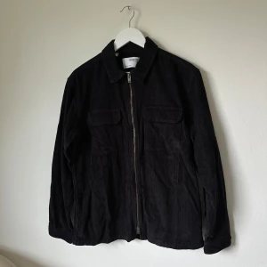 Svart manchesterjacka från Selected Homme - Snygg svart manchesterjacka från Selected Homme med klassisk krage, två bröstfickor och dragkedja framtill. Jackan är tillverkad i mjuk bomull och har en rak passform med långa ärmar. Perfekt för dig som gillar stilren och enkel design. 2N9