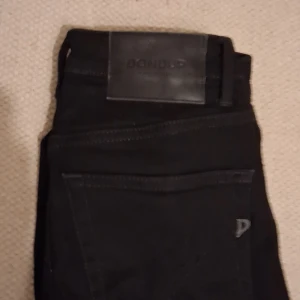 Svarta jeans från Dondup - Snygga svarta jeans från Dondup, tillverkade i Italien. Klassisk femficksmodell med svarta sömmar och diskret Dondup-logga på bakfickan. Jeansen har en rak passform och är gjorda i ett mjukt bomullsmaterial med lite stretch för extra komfort. Storlek w26