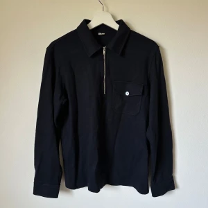 Mörkblå långärmad tröja från Arket - Mörkblå långärmad tröja från Arket med klassisk krage, bröstficka med knapp och half zip-dragkedja framtill. Tröjan har en clean look och är tillverkad i en mjuk materialmix som känns skön mot huden. 2K2