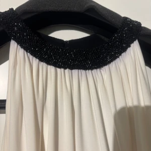 Svartvit långklänning med pärldetaljer - Elegant långklänning i svart och vitt med ärmlös design. Överdelen är vit med vackert draperat tyg och en hög halsringning dekorerad med svarta pärlor. Kjolen är svart och faller rakt ner. Perfekt för dig som vill sticka ut med en stilren och sofistikerad look.