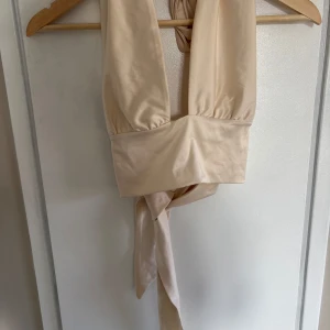 Beige omlottopp från Gina Tricot - Säljer en topp från  Gina Tricot i glansigt satinliknande material. Toppen har breda axelband, djup v-ringning och knytdetaljer både fram och bak för en snygg och justerbar passform. Perfekt för dig som gillar trendiga och stilrena plagg.