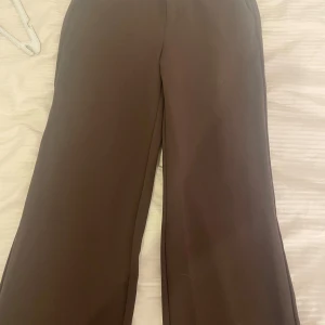 Bruna kostymbyxor från Vero Moda - Snygga bruna kostymbyxor från Vero Moda med raka, vida ben och hällor för bälte. Byxorna har klassisk passform och är perfekta för en stilren look. Tillverkade i ett mjukt material som känns bekvämt hela dagen.