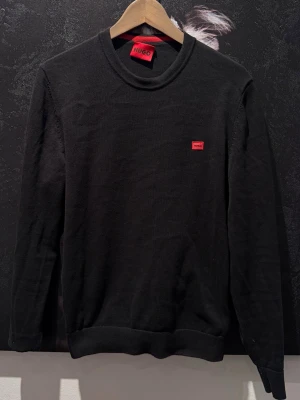 Svart stickad tröja från Hugo - Snygg svart stickad tröja från Hugo med rund halsringning och en liten röd logotyp på bröstet. Tröjan har ribbade muddar vid ärmslut och nederkant. Perfekt för dig som gillar stilrena och klassiska plagg med en modern touch.