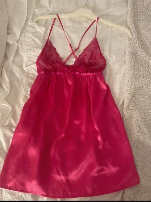 Rosa satin nattlinne med spetsdetaljer - Snyggt nattlinne i starkt rosa satin med tunna axelband och spets upptill. Axelbanden är korsade i ryggen och kuporna har fina spetsdetaljer. Materialet är glansigt och mjukt, perfekt för en bekväm kväll hemma. Ingen aning vilket märke de är ifrån 