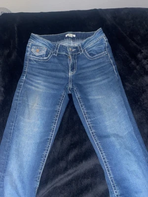 Blå bootcut jeans med vita sömmar - Säljer ett par klassiska blå bootcut jeans med snygga vita kontrastsömmar och fem fickor. Jeansen har en låg midja och är tillverkade i mjukt denimtyg som ger en avslappnad vibe. Perfekta för dig som gillar en tidlös look.