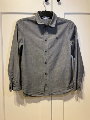 Grå skjorta med knappar från H&M - Stilren grå skjorta från H&M med klassisk krage och svarta knappar framtill. Skjortan har lång ärm och en liten broderad logga på bröstet. Tillverkad i mjuk bomull som känns skön mot huden. Perfekt för en clean och enkel look.