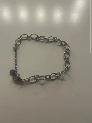 Silverfärgat kedjearmband med berlock - Stilrent armband i silverfärgad metall med ovala länkar ni och en liten rund berlock. Kedjan har en lätt vriden design som ger extra detalj. Perfekt för dig som gillar minimalistiska accessoarer med en twist.