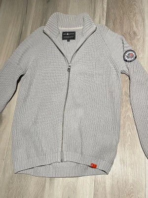 Ljusgrå stickad ziptröja Sail Racing - Ljusgrå stickad tröja från Sail Racing med dragkedja framtill, hög ribbad krage och patch på ena ärmen. Tröjan har en grovstickad struktur och ribbade muddar. Perfekt för lager-på-lager och snygg till jeans.