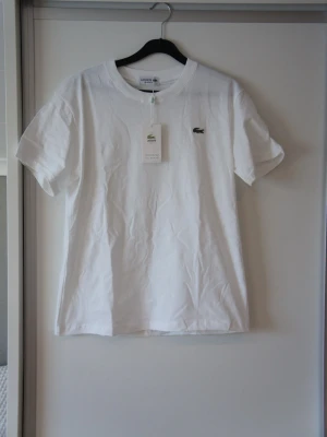 Vit t-shirt från Lacoste - Klassisk vit t-shirt från Lacoste med den ikoniska gröna krokodilloggan broderad på bröstet. Tillverkad i mjuk bomull och har en normal passform med korta ärmar och rund halsringning. Perfekt basplagg för en clean och stilren look.