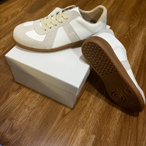 Maison Margiela vita sneakers - Stilrena sneakers från Maison Margiela i vitt och beige med detaljer i mocka och skinn. Klassisk låg modell med rund tå, snörning och platt sula. Kommer med originalkartong och dustbag.