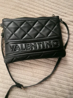 Svart quiltad axelväska Valentino - Snygg svart axelväska från Valentino med quiltad design och justerbar axelrem. Väskan har ett stort VALENTINO-tryck framtill och stängs med dragkedja. Tillverkad i skinnimitation och har en stilren, modern look.