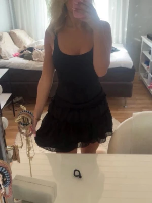 Svart volangkjol med resårmidja - Supersöt svart kortkjol med volanger och smockad resår i midjan. Kjolen har ett lätt och luftigt tyg som ger en flowy känsla. Perfekt att styla med en enkel topp eller stickad tröja.