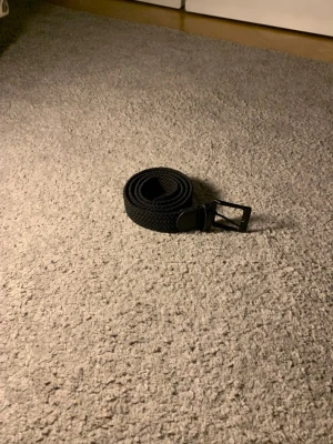 Svart flätat bälte med metallspänne - Snyggt svart flätat bälte med klassiskt fyrkantigt metallspänne och detaljer i svart läder. Perfekt accessoar för att lyfta din outfit och passar till både jeans och chinos. Bältet har en stilren design och är lätt att matcha med olika stilar.