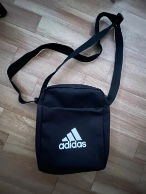 Svart axelväska från Adidas - Snygg svart axelväska från Adidas med justerbar rem och vit logga framtill. Väskan har dragkedja upptill och ett extra fack med dragkedja på framsidan. Perfekt storlek för det viktigaste och enkel att bära crossbody.