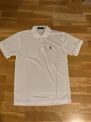 Vit pikétröja från Polo Ralph Lauren - Klassisk vit pikétröja från Polo Ralph Lauren med korta ärmar och krage. Tröjan har två knappar framtill och den ikoniska mörkblå logotypen broderad på bröstet. Tillverkad i bomull för en skön och luftig känsla.