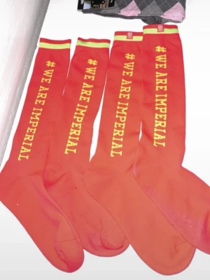Röda knästrumpor #WE ARE IMPERIAL - Säljer fyra par röda knästrumpor från Imperial med texten '#WE ARE IMPERIAL' i gult längs benet. Strumporna har en grön och gul rand upptill och är perfekta för dig som vill sticka ut. Materialet känns mjukt och stretchigt. Även finns det ett par vanliga rid strumpor.