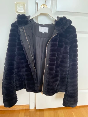 Svart pälsjacka med huva från Vila - Mysig svart pälsjacka från Vila med huva och dragkedja framtill. Jackan har ett fluffigt, randigt mönster i fuskpäls och är perfekt för kalla dagar. Passformen är normal och jackan är helsvart vilket gör den enkel att matcha.