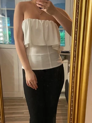 Vit axelbandslös bandeautopp från Zara - Säljer en snygg vit bandeautopp från zara. Toppen är axelbandslös med volangdetalj upptill och har en croppad passform. Ny skick utan lapp, använd en gång inga defekter alls.