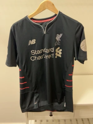 Liverpool bortatröja Mané 19 - Svart Liverpool FC bortatröja från New Balance med tryck 'Mané 19' på ryggen. Tröjan har vita och röda detaljer, klubbmärke och sponsorlogga på bröstet. Perfekt för fotbollsfans som vill visa sitt stöd för laget.