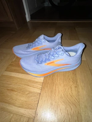 Brooks löparskor Ghost 17 - Snygga löparskor från Brooks i lila meshmaterial med orange detaljer och logga på sidan. Skorna har en sportig siluett, tjock sula för bra dämpning och ventilerande ovandel. Perfekta för dig som vill ha stilrena och lätta skor till löpningen.