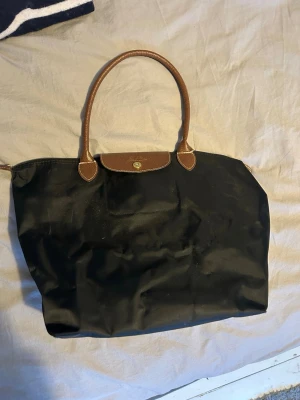 Svart handväska från Longchamp - Klassisk svart handväska från Longchamp med bruna handtag och lock i skinn. Väskan har en enkel och stilren design med guldfärgad knappdetalj. Mått: 31x30x19cm Handtagets höjd: 25cm. Trasig i ena handtaget, går att sy fast för den som har kapaciteten. 1 599kr ny. 