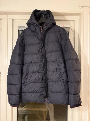 Marinblå dunjacka från Herno med huva - Snygg marinblå dunjacka från Herno med huva och dragkedja framtill. Jackan har quiltad design, två sidofickor och hög krage för extra värme. Perfekt för kalla dagar och enkel att matcha med olika outfits.