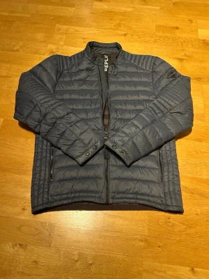 Mörkblå dunjacka från Replay - Snygg mörkblå pufferjacka från Replay med quiltad design och hög krage. Jackan har dragkedja framtill, två sidofickor med dragkedja och Replay-logga på kragen. Perfekt för kyliga dagar och enkel att matcha till olika outfits. Enda defekten är att Y ett som är på dragkedjan är borta, alltså klisterlappen som sitter ovanpå men man ser fortfarande att det står REPLAY. Andra är att man kan se lite slitage på knappar men då ska man kolla noga. MVH Liam/Brakansgarderob