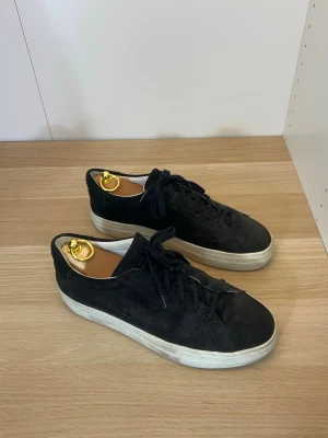 A Day’s March Skor | 42 - Stilrena svarta sneakers i mocka från A Day’s March med klassisk låg profil och vita gummisulor. Skorna har svarta skosnören och en enkel, tidlös design som passar till det mesta. Perfekta för dig som gillar en clean och avslappnad look.