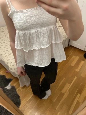 Vit volangtop med smock - Säljer en söt vit topp med smock upptill och dubbla volanger nedtill. Tunna axelband och luftig, krinklad bomullskänsla gör den perfekt för sommaren. Passar dig som gillar romantisk och avslappnad stil.