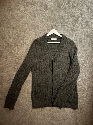 Stickad Kofta - Jag säljer en kabelstickad kofta i grå färg. En riktigt bekväm sådan med fem knappar, vilket hjälper dig att kunna justera hur du vill ha det! Tröjan är från Bläck i storlek Large. Tröjan är gjord av 50% bomull och 50% ull.
