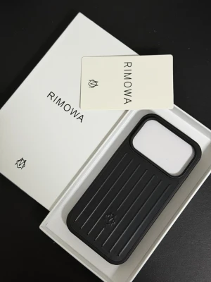 Svart iPhone 17 Pro Rimowa mobilskal  - Stilrent svart mobilskal från Rimowa med räfflad design och diskret logotyp. Skalet är tillverkat i slitstarkt material och har en exakt passform för iPhone. Perfekt för dig som vill ha ett exklusivt och skyddande skal med modern känsla.