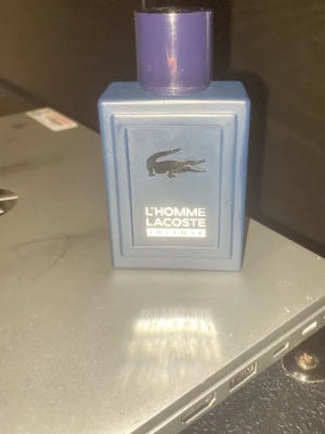 Lacoste L'Homme Intense parfym - Säljer en Lacoste L'Homme Intense parfym i en stilren, fyrkantig blå flaska med lila kork och den klassiska krokodilloggan framtill. Flaskan är tillverkad i glas och har en modern, maskulin design som sticker ut i badrumshyllan. Nästan aldrig använd tagit kanske 10-20 sprutor av den