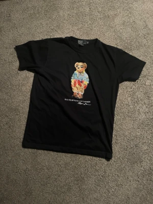 Mörkblå Polo Bear t-shirt från Ralph Lauren - Mörkblå t-shirt från Polo Ralph Lauren med ikoniskt Polo Bear-tryck på bröstet. Klassisk passform och rund halsringning. Tillverkad i mjuk bomull, perfekt för en avslappnad och stilren look, passar också S.
